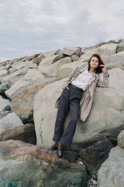 Bulutlu bir havada Rocky Beach 'te büyük kayalara yaslanan sıradan bir genç kadın. Doğal duruş ve açık hava gösterimi rahat stil ve doğa kavramı ile bağlantı.