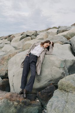 Sıradan giyinmiş bir kadın kayalık sahilde duruyor, elleri saçlarında taşa yaslanıyor. Bulutlu hava ve doğal kayalar açık hava atmosferini ve sessiz tefekküre vurgu yapıyor..