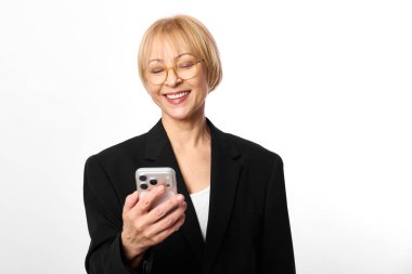 Kısa sarı saçlı ve gözlüklü mutlu orta yaşlı kadın akıllı telefonu tutuyor ve ekrana bakarken gülümsüyor. Siyah ceket ve beyaz arka plan temiz ve modern bir atmosfer yaratır..