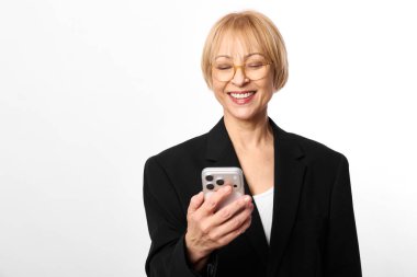 Gözlüklü ve siyah ceketli mutlu orta yaşlı kadın akıllı telefon tutuyor ve beyaz arka plana bakarken gülümsüyor. Dijital iletişim, teknoloji, sosyal medya kavramı ve