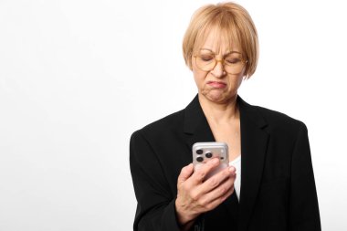 Hüsrana uğramış, orta yaşlı, gözlüklü ve siyah ceketli bir kadın, beyaz arka planda hoşnutsuz bir ifadeyle akıllı telefon tutuyor. Teknoloji kullanımı, iletişim sorunları ve olumsuz duygular kavramı.