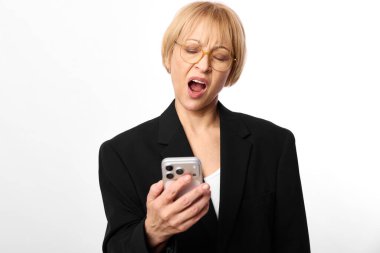 Gözlüklü, siyah ceketli, sinirli olgun bir kadın elinde akıllı bir telefonla hayal kırıklığını ve hoşnutsuzluğunu dile getiriyor. Beyaz arka plan stüdyo çekimi. İletişim sorunları ve stres kavramı.