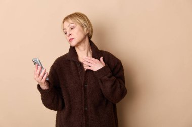 Kahverengi ceket giyen düşünceli orta yaşlı bir kadın, arka plandaki bej rengi gözlerden uzağa bakan akıllı bir telefon tutuyor. Teknoloji, iletişim ve modern yaşam tarzı kavramı.