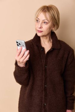 Kahverengi hırkalı sarışın kadın odaklanmış bir ifadeyle akıllı telefona bakıyor. Nötr bej arka plan gelişigüzel stil ve dijital cihaz kullanımını vurgular. Modern iletişim ve yaşam tarzı kavramı.