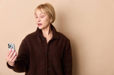 Kahverengi ceketli sarışın kadın akıllı telefonu tutuyor ve şaşkınlıkla tepki veriyor. Minimalist bej stüdyo ayarları yüz ifadesini ve gündelik sonbahar kıyafetlerini ön plana çıkarıyor.