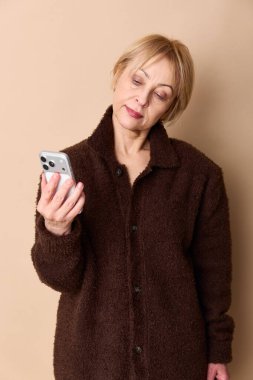 Koyu kahverengi kazak giyen yaşlı bir kadın akıllı telefonu elinde tutuyor ve yorgunluktan yere bakıyor. İzole edilmiş bej arkaplan iletişim, teknoloji ve olgun yaş temalarını vurguluyor.