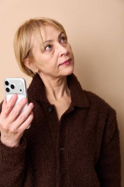 Kahverengi ceket giyen düşünceli orta yaşlı kadın akıllı telefonu tutuyor ve bej arka planda yukarı bakıyor. Teknoloji kullanımı, iletişim, düşünce ve modern yaşam tarzı kavramı.