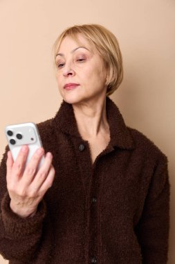 Kahverengi desenli ceketli sarışın, orta yaşlı kadın akıllı telefon ekranına odaklanmış. Bej arka planı ve yumuşak doğal ışığı olan kapalı bir portre. İletişim ve teknoloji kavramı.