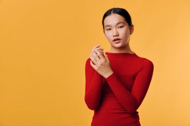 Kırmızı uzun kollu bluz giyen genç bir kadın kapalı mekanda poz veriyor, elleri sımsıkı bağlı ve sakin bir yüzü var. Moda veya duygu konsepti için parlak turuncu arkaplan içeren stüdyo portresi.