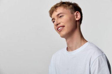 Gündelik sahnede beyaz tişörtlü, yumuşak nötr bir zemine karşı gevşek bir duruş takınan genç bir adam markalaşma, reklam ve modern yaşam tarzı görselleri için yaklaşılabilir enerji aktarıyor.