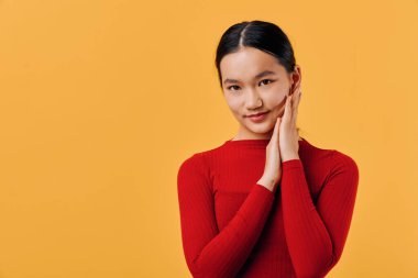 Yüzüne yakın elleri olan kırmızı elbiseli bir kadın kameraya bakıyor. Temiz ciltli ve pürüzsüz siyah saçlarıyla gülümsüyor. Modern stüdyoda parlak turuncu duvarın karşısında duruyor..