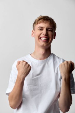 Adam zafer ve motivasyon için yumruklarını kaldırır, tişörtle geniş bir şekilde gülümser, özgüveni ve canlılığı yansıtır, reklam, kampanya ya da stüdyo ışıklandırması ve pozitif markalaşma için uygundur.