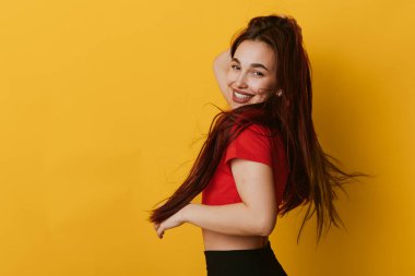 Kırmızı bluzlu, uzun saçlı, gülümseyen genç bir kadın. Parlak sarı arkaplanda poz veriyor. Gündelik tarzı, neşeli havası ve yaşam tarzı ya da moda konsepti için dinamik duruşu olan kadın portresi.