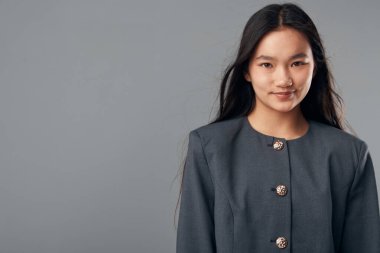 Hafif bir gülümsemeyle poz veren, koyu renk ceketli, düğmeli, gri arka planda kameraya bakan, kendine güvenen, kendine güvenen, güzel ve moda portresi olan genç bir kadın..