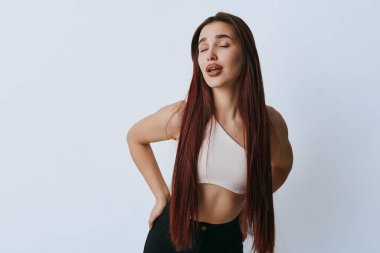 Uzun örgülü, sırtına dokunan, gözleri kapalı genç bir kadın sırt ağrısı ya da tarafsız arka planda giydiği sıradan kıyafetlerle kas gerilimi gösteriyor..