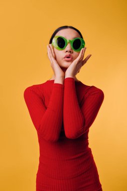 Parlak yeşil güneş gözlüklü ve kırmızı uzun kollu kadın model sarı arka fonun önünde duruyor, neşeli ve hayret verici bir yüz ifadesiyle yanakların yanında el ele tutuşuyor..