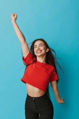 Kırmızı gömlekli, siyah pantolonlu, uzun saçlı, gülümseyen genç bir kadın mavi duvara yaslanıyor. Enerjik bir kadın mutluluk ve hareketini kapalı gözlerle ve neşeli bir ifadeyle gösteriyor..