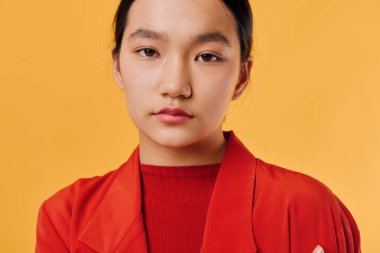 Kırmızı kıyafetli, turuncu arka planda duran Asyalı kadın. Makyajı az, rahat tarzı ve stüdyo ışığında sade saçı olan kendine güvenen bir kadın portresi..