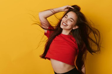 Smiling young woman in red shirt with flowing hair and closed eyes on yellow backdrop. Portrait of joyful female model with casual style and relaxed expression in bright setting.