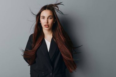 Kendine güvenen, uzun düz saçlı, siyah ceketli, beyaz bluzlu genç bir kadının portresi gri duvarın önünde duruyor. Tarz, zarafet ve modern görünüş için..