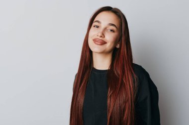 Confident young woman with straight red hair wearing black top posing on plain light gray background. Female portrait with subtle smile and natural look in studio setting.