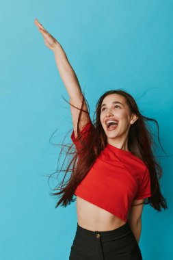 Kırmızı gömlekli, uzun dalgalı saçlı, gülümseyen kız mavi arka planda el kaldırıyor. Genç, neşeli bir yüz ifadesi, günlük kıyafetler ve parlak iç mekanlarda kaygısız bir tavır..