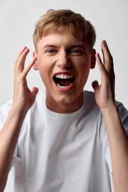 Neşeli patlamalar ve yüksek sesli tepkiler, gülümseyen bir adamın bağırışları, ellerinin başının yanında olması, beyaz tişört, stüdyo düzeni, yoğun enerji ve anlamlı an.