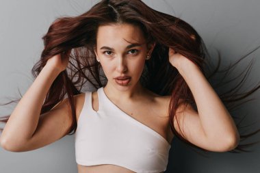 Beyaz ekin bluzlu kameraya bakarken saçı kabarık bir kadın model. Stüdyo portresi. Doğal makyajlı ve bakışları gri arka plana odaklanmış genç bir kadın..