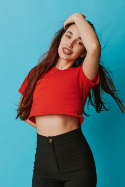 Confident young woman with long hair in red crop top and black pants posing on blue background. Beauty, fashion, casual clothing and studio portrait of smiling female model.