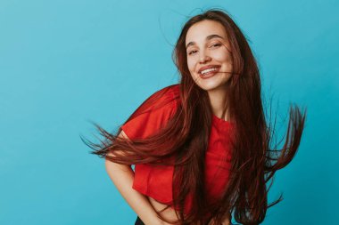 Kırmızı gömlek giyen, dalgalı saçlı, gülümseyen genç bir kadının portresi. Mavi arka plan, günlük moda, neşeli ruh hali, ve parlak stüdyo ortamında canlı saç hareketi..