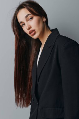 Young woman with sleek brown hair and subtle makeup dressed in black jacket. Studio portrait of attractive female with serious look and smooth hair on plain gray backdrop.