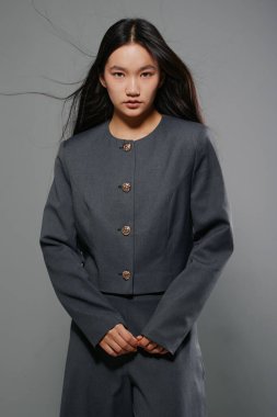 Gri elbiseli, siyah saçlı kadın doğrudan kameraya bakıyor. Tarafsız gri arkaplanlı stüdyo çekimi, şık giyinme ve moda veya iş konsepti için doğal makyaj.