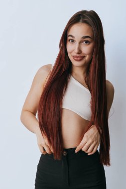 Confident young woman with long brown hair dressed in white sleeveless top and black pants standing with smile and casual pose on plain light background indoors.