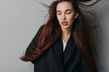 Siyah ceketli, uzun saçlı, gözleri kapalı gri arka planda poz veren genç bir kadın. Pürüzsüz ciltli yakın plan portre ve tarz, moda veya güzellik çekimi için gevşek ifade.