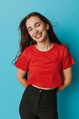 Kırmızı gömlekli ve siyah pantolonlu mutlu bir kadın. Gözleri kapalı poz veriyor ve elleri mavi arka planda, uzun koyu renk saçlar ve neşeli bir gülümseme sergiliyor..