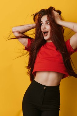 Happy young woman with red top and long hair standing on yellow background. Energetic pose, smiling face, casual clothing and positive emotions in colorful studio shot.