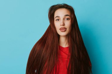 Uzun kahverengi saçlı ve kırmızı bluzlu genç bir kadın portresi. Mavi duvarın önünde duruyor. Stüdyoda güzellik ve moda konsepti için sakin bir yüz ve doğal makyaj..