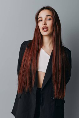 Portrait of fashionable young woman with long hair dressed in black jacket and white crop top posing on gray studio background with relaxed posture and direct gaze.