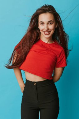 Smiling female model with loose hair dressed in red crop top and black trousers standing with hands behind back on blue backdrop. Youthful and relaxed mood.
