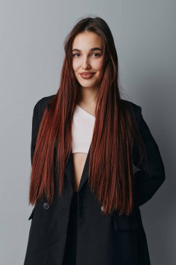 Uzun kahverengi saçlı, siyah ceketli, beyaz gömlekli, elleri arkada, stüdyo ışıklarında gri arka planda poz veren kendine güvenen genç bir kadının portresi..