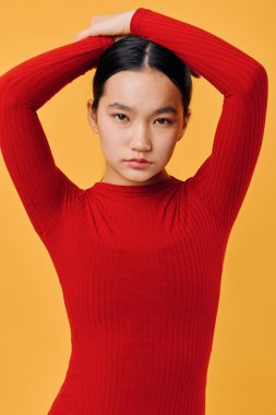 Portrait of confident young woman in red long sleeve sweater, hair tied back with hands resting on head, isolated on bright yellow background with focused expression.