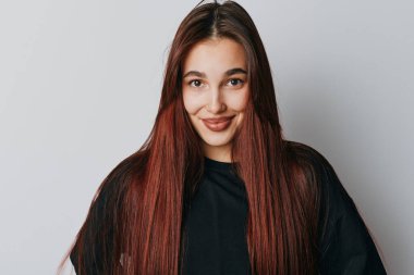 Smiling young woman with long brown hair wearing black top posing in studio. Clean white background, natural look and friendly face for personal or fashion portrait.