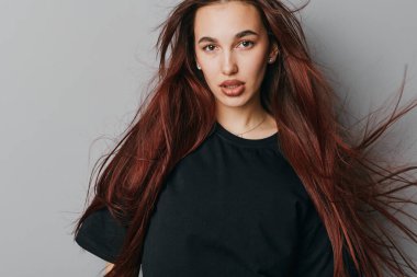 Kendinden emin, kırmızı saçlı ve siyah tişörtlü bir kadının portresi gri arka planda poz veriyor, doğal güzelliği ve en az aksesuarla direkt bakışları gösteriyor..