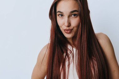 Uzun kahverengi saçlı, beyaz arka planda rahat duran gülümseyen genç bir kadının portresi. Sağlıklı saçlı ve kendine güveni olan bir kadın güzellik konsepti için yakın plan çekimde..
