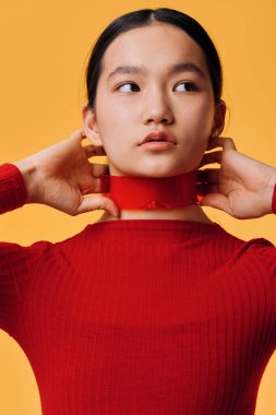 Female model with tied hair wearing red long sleeve top and red neck decoration posing in studio. Young Asian woman with clear skin and focused look on yellow backdrop.