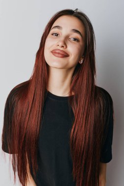 Smiling young woman with straight red hair wearing black t shirt and small earrings posing in front of white wall. Closeup portrait of confident female with natural look and relaxed expression.