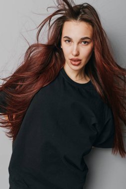 Siyah tişörtlü, bol saçlı genç bir kadın içeride poz veriyor. Doğal görünümlü ve ciddi yüzlü bir kadın model. Tarafsız bir arkaplan önünde. Yaşam tarzı ya da güzellik kavramı için..