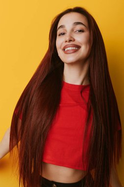 Smiling young woman with long brown hair wearing red crop top posing confidently on yellow background. Female portrait with natural expression and casual style in studio.