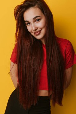 Smiling young woman with long brown hair in red crop top standing in front of yellow wall. Female model posing confidently with casual outfit and natural look for lifestyle or fashion concept.