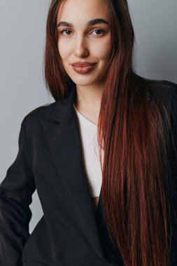 Uzun kahverengi saçlı, siyah ceketli ve beyaz gömlekli, profesyonel ya da sıradan fotoğraf çekimi için gri duvarın önünde duran kendine güvenen genç bir kadın..